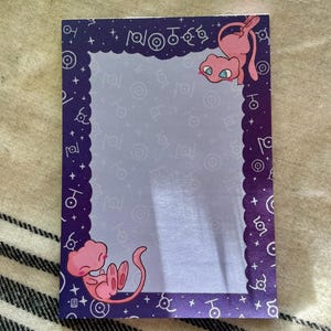 Puede incluir: Un bloc de notas rectangular con un borde morado con borde festoneado y estrellas blancas. El bloc de notas tiene un interior lila claro con un patr&oacute;n repetido de s&iacute;mbolos blancos. Se representan dos criaturas de dibujos animados rosas.