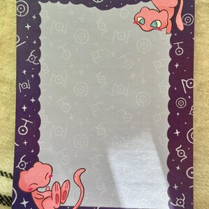 Puede incluir: Un bloc de notas rectangular con un borde morado con borde festoneado y s&iacute;mbolos blancos. Se representan dos criaturas de dibujos animados rosas, una colgada boca abajo y la otra acurrucada. El fondo es gris claro con s&iacute;mbolos repetidos.