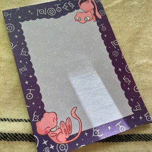 Puede incluir: Bloc de notas rectangular con borde morado e interior morado claro. El borde presenta un borde festoneado y s&iacute;mbolos blancos. Se representan dos criaturas de dibujos animados rosas, una en la parte superior y otra en la inferior.