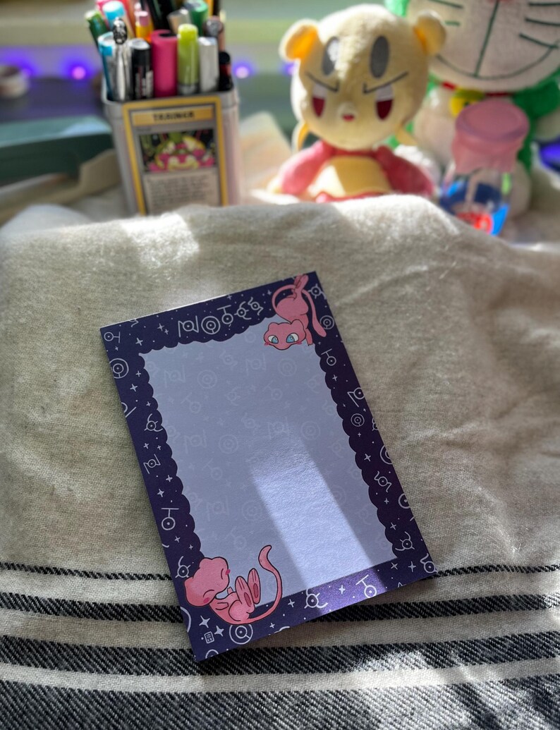 Puede incluir: Un bloc de notas rectangular con un borde morado y un interior azul claro. El borde presenta un borde festoneado y peque&ntilde;as estrellas blancas. Dos personajes de dibujos animados rosas est&aacute;n en el bloc de notas. El bloc de notas est&aacute; sobre una superficie rayada blanca y gris.