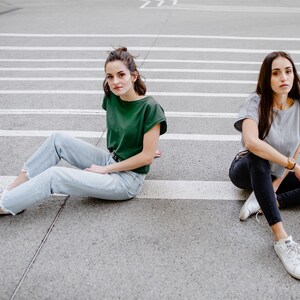 May include: Two people are sitting on a gray concrete surface with white lines. The person on the left is wearing a green t-shirt, blue jeans, and white sneakers. The person on the right is wearing a gray t-shirt, black jeans, and white sneakers.