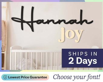 May include: A wall-mounted sign featuring the name Hannah in a black cursive script font positioned above the word joy in a light beige serif font. The letters are mounted on a plain wall surface above a white crib with vertical slats.