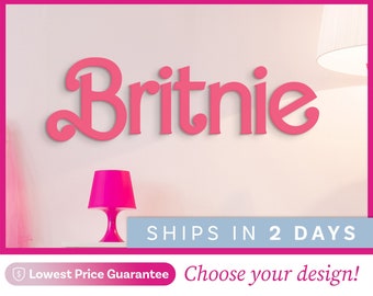 May include: A wall-mounted sign featuring the name Britnie in a bold, pink, cursive-style font. The sign is displayed against a light-colored wall above a bright pink table lamp. The text 2day decor is visible in the top left corner of the image.