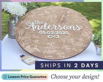 May include: A round wooden guest book sign featuring the white text "the Andersons" and "05.02.2020" with an infinity symbol below. The surface is covered in handwritten signatures and messages in white ink, displayed on a white table.