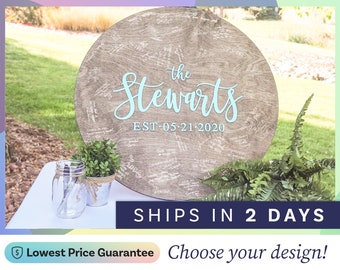 May include: A round wooden guest book sign with a light brown wood grain finish, featuring the text "the Stewarts" in light blue script and "EST 05-21-2020" in light blue block letters, surrounded by numerous handwritten signatures in white ink.