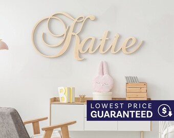 May include: A light-colored wooden wall sign featuring the name Katie in a cursive script font. The letters are connected in a flowing style, mounted on a plain wall above a white cabinet with various decorative items including a pink plush toy.