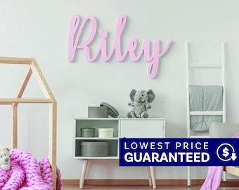 May include: A light pink wall-mounted sign featuring the name Riley in a cursive script font with a small heart above the letter i. The sign is displayed on a plain wall above a white storage cabinet containing various round boxes and a plush elephant.