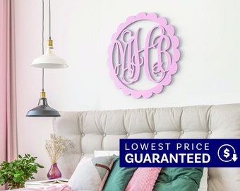 May include: A circular light pink wall decoration featuring a scalloped edge and an intricate, interconnected monogram of the letters M, H, and B in a cursive script font, mounted centrally on a plain white wall above a tufted headboard.