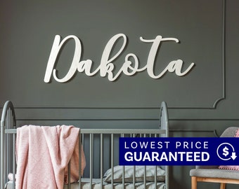 May include: A white wall-mounted sign featuring the name Dakota in a cursive script font. The letters are connected and have a smooth finish. The sign is displayed horizontally against a dark gray wall above a crib with a pink blanket.