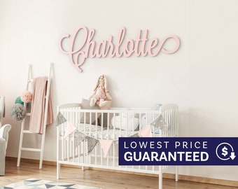 May include: A light pink wooden wall sign featuring the name Charlotte in a cursive script font. The letters are connected in a single piece, displaying a smooth matte finish and mounted horizontally on a plain white wall above a white crib.