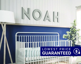 May include: Large gray capital letters spelling the name NOAH mounted on a white paneled wall above a white metal crib. The crib features a thin vertical bar design and a light blue and white patterned mattress cover in a nursery setting.