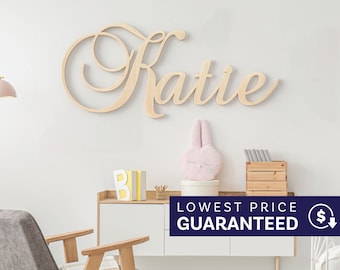 May include: A light-colored wooden wall sign featuring the name Katie in a cursive script font. The letters are connected in a flowing style, mounted on a plain wall above a white cabinet with various decorative items including a pink plush toy.