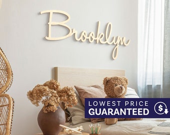 May include: A light-colored wooden wall sign featuring the name Brooklyn in a cursive script font. The letters are connected and mounted horizontally on a wall above a bed, displaying a smooth, natural wood finish with a clean, simple design.