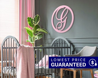 May include: A circular wall sign featuring a large, stylized letter G in a soft pink finish. The letter is enclosed within a matching pink circular frame, mounted on a dark gray wall above a gray crib and a gray upholstered chair.