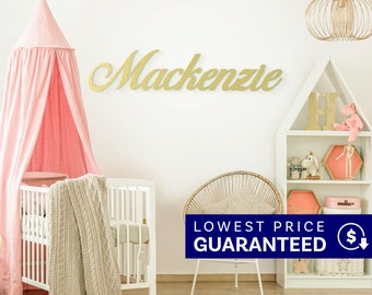 May include: A gold-colored wall-mounted sign featuring the name Mackenzie in a cursive script font. The sign is displayed on a white wall above a white crib with a pink canopy, a rattan chair, and a white house-shaped shelving unit with toys.