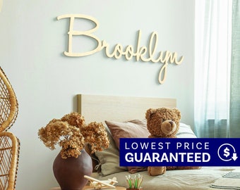 May include: A light-colored wooden wall sign featuring the name Brooklyn in a cursive script font. The letters are connected and mounted horizontally on a plain wall above a bed, displaying a smooth, natural wood finish with clean edges.