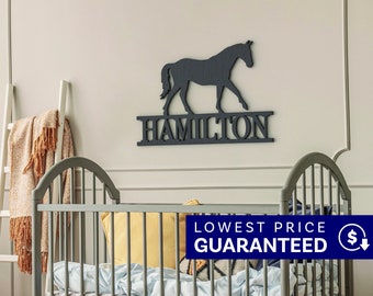 May include: A dark gray wall-mounted sign featuring a silhouette of a horse standing above the name HAMILTON in capital letters. The sign has a textured finish and is mounted on a plain wall above a gray crib with bedding and pillows.