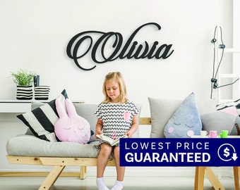 May include: A black wall-mounted sign featuring the name Olivia in a cursive script font. The lettering is constructed from a solid material and is mounted horizontally on a plain white wall above a light-colored wooden sofa with cushions.