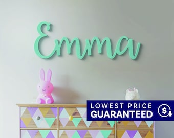 May include: A mint green wall-mounted sign featuring the name Emma in a cursive script font. Below the sign is a wooden dresser with a geometric pattern of triangles in shades of purple, yellow, and green, topped with a pink rabbit lamp.