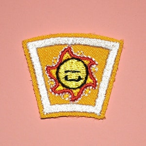 以下が含まれることがあります： 笑顔の太陽のデザインが施された刺繍パッチ。パッチは黄色の背景に白い縁取りがあり、中央に赤と黄色の太陽のグラフィックがあります。パッチは台形です。