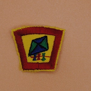 May include: A red, yellow, and blue embroidered patch with a kite design. The patch is shaped like a pentagon and has a yellow background with a red border.