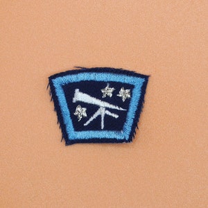 May include: Embroidered patch with a blue border and a dark blue interior. The patch features a white telescope and three white star-like shapes. The patch is on a peach-colored background.