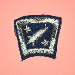 May include: A dark blue embroidered patch with a white border. The patch features a white diagonal streak and two white stars on a dark blue background. The patch is on a pink background.
