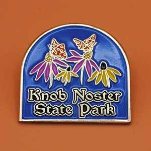 May include: Blue and gold enamel pin with a purple, yellow, and orange flower design and three butterflies. The text "Knob Noster State Park" is printed in black on the pin.