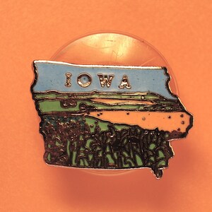 以下が含まれることがあります： アイオワ州の形をした青と緑のエナメルピン。ピンには白い文字で「IOWA」と書いてあります。