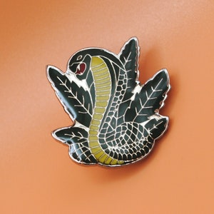 May include: A detailed enamel pin featuring a cobra with a raised hood. The snake is depicted in black, yellow, and white, with a red tongue. The pin is set against a backdrop of stylized black leaves and has a silver border.