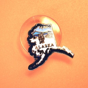May include: A blue and white enamel pin shaped like the state of Alaska with the word "ALASKA" printed in white on the pin. The pin is attached to a clear plastic backing.