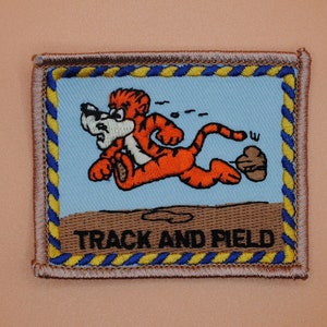 May include: A brown and blue embroidered patch with a cartoon tiger running. The patch has the text "TRACK AND FIELD" in black.