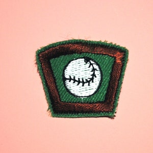 May include: A green and brown embroidered patch with a white baseball inside. The baseball has black stitching.