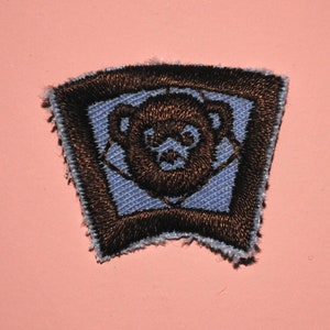 May include: A blue and brown embroidered patch with a bear face in the center.