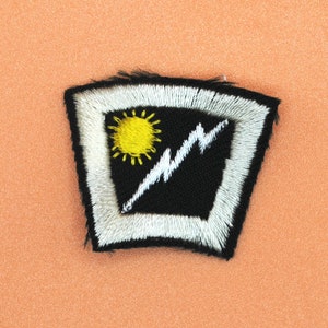 以下が含まれることがあります： 白い稲妻と黄色の太陽が刺繍された黒と白のワッペン。ワッペンは白い縁取りで、湾曲した形をしています。
