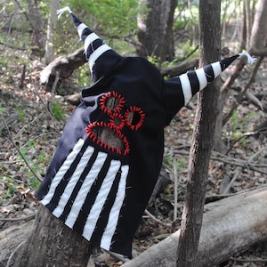 May include: A black hooded costume with white stripes and pointed horns. The costume has red stitching and is resting on a tree trunk in a forest setting. The design is reminiscent of a whimsical, handcrafted character.