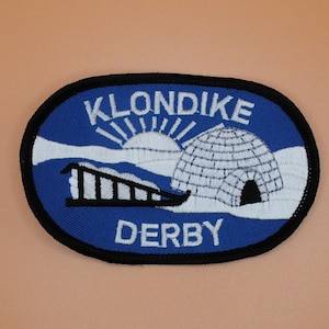 May include: Oval patch with a blue background and black border. The patch features the words "KLONDIKE DERBY" in white, along with an igloo, a sun, and a sled. The patch is likely a souvenir or emblem.