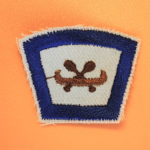 May include: Embroidered patch with a white center featuring a brown canoe and crossed oars. The patch has a blue border and is set against an orange background. The patch is likely for a uniform or clothing.