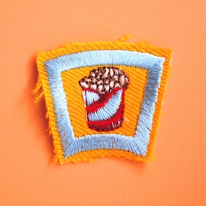 May include: A yellow and blue embroidered patch with a red and white striped popcorn bucket.