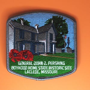 May include: A rectangular embroidered patch with rounded corners featuring a house, a statue, and two red poppies. The text reads: GENERAL JOHN J. PERSHING BOYHOOD HOME STATE HISTORIC SITE LACLEDE, MISSOURI. The patch has a grey border.