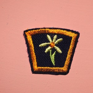 May include: A dark blue embroidered patch with a yellow flower design, surrounded by an orange border. The patch is shaped like a trapezoid.