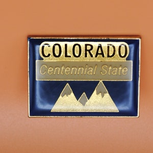 May include: A gold-colored enamel pin with a blue background. The pin features the word "COLORADO" in gold lettering and the text "Centennial State" in white lettering. The pin also features a gold-colored mountain range.
