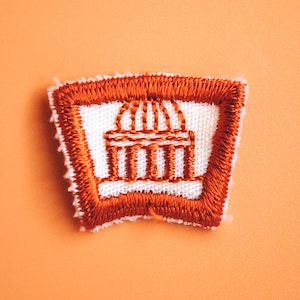 May include: A brown embroidered patch with a white building design. The building has a dome and columns. The patch is a Girl Scout badge.
