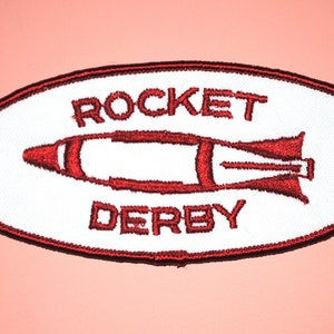 May include: White oval embroidered patch with red stitching. The patch features a red rocket ship and the text "ROCKET DERBY".