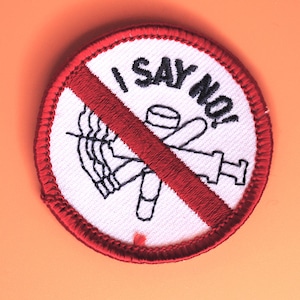 May include: A circular patch with a white background and a red border. The patch features a red diagonal line through a black and white illustration of a syringe, a pill, and a cigarette. The text "I SAY NO!" is embroidered in black.