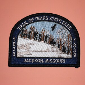 May include: A blue and white embroidered patch with the text "Trail of Tears State Park 1838 Jackson, Missouri 1838". The patch depicts a group of people walking through a snowy forest with a bird flying overhead.