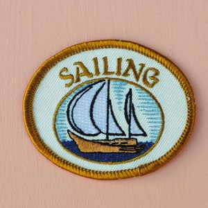 以下が含まれることがあります： 金色と白の刺繍が施されたパッチで、金色の文字で「SAILING」と書いてあり、青と白の帆船が描かれています。船体は茶色です。