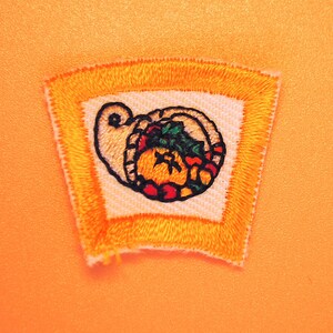May include: A yellow embroidered patch with a white border and a colorful image of a cornucopia filled with fruit and vegetables.