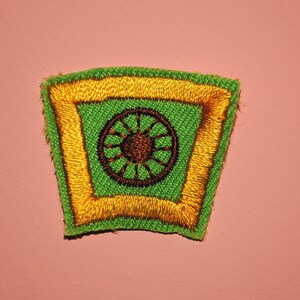May include: Embroidered patch featuring a green background with a gold border. The patch has a brown wheel design in the center. The patch is a trapezoid shape with a pink background.