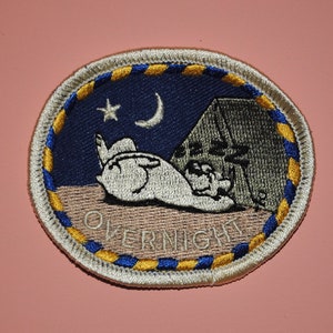 May include: A white and blue embroidered patch with a crescent moon, a star, and a sleeping bunny in front of a tent. The text "OVERNIGHT" is embroidered in white on a beige background.